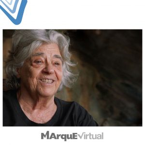 Fotografia de uma mulher idosa com cabelos grisalhos e ondulados, vestindo uma camiseta preta. Ela está sorrindo de maneira serena e acolhedora. O fundo é desfocado, com tons terrosos, sugerindo um ambiente natural ou rústico. No topo da imagem há um logotipo com formas em azul e branco. Na parte inferior, aparece a inscrição “MArqueEVirtual” em letras estilizadas.